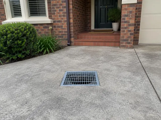 Concrete driveway with a square outdoor gully grate beside brick steps and a front door in Killara